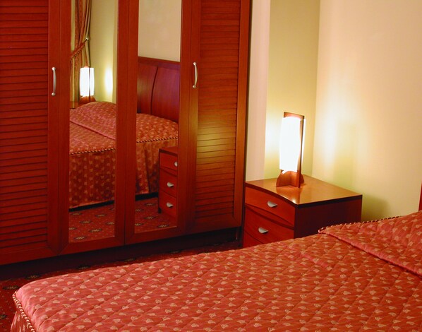 Habitación de hotel con cama, mesita de noche con lámpara, armario con espejo y piso alfombrado.