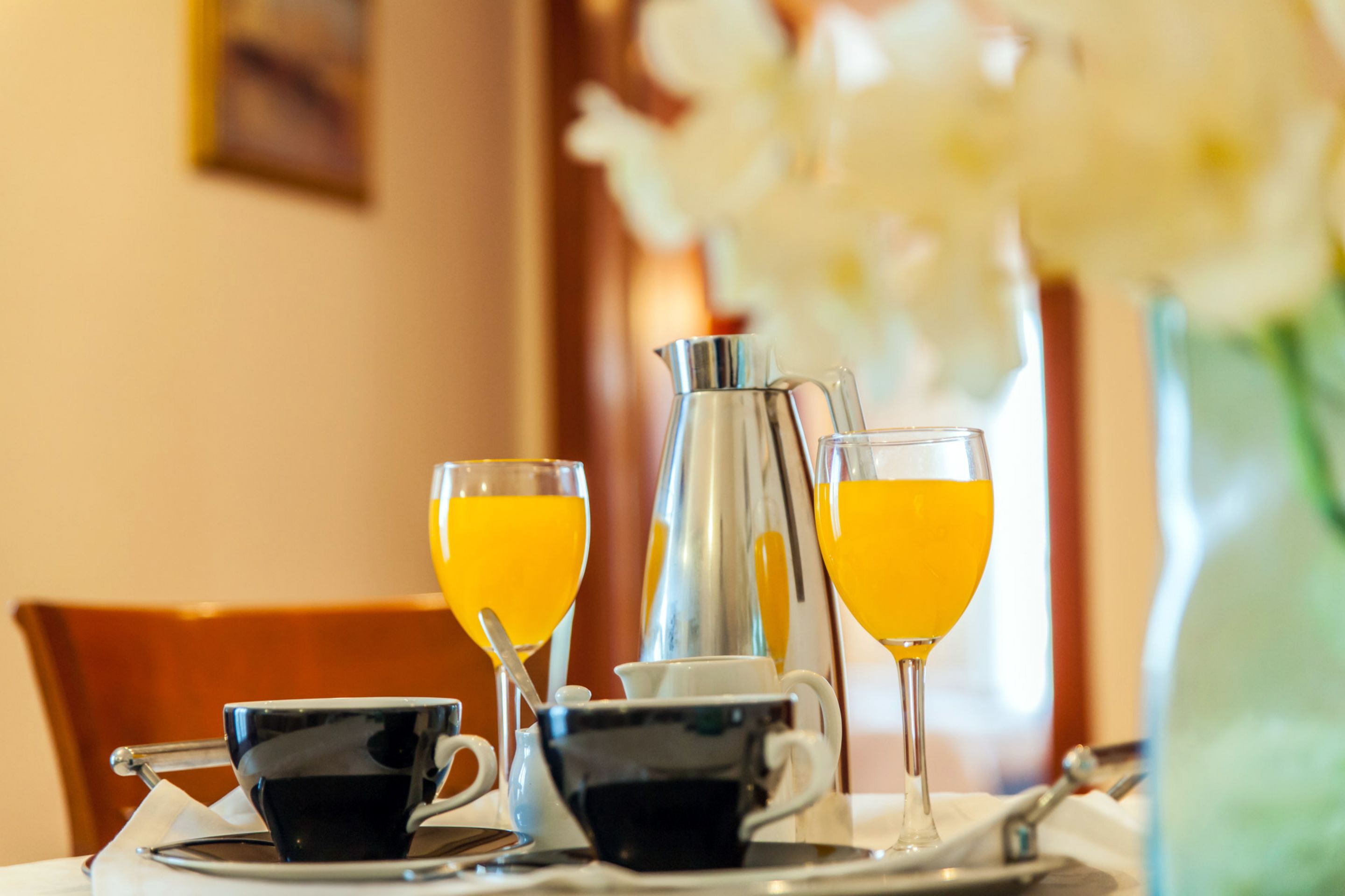 Una escena de desayuno con dos tazas de café, dos vasos de jugo de naranja, una jarra de plata y un florero con flores blancas.