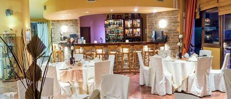 Un restaurante con mesas redondas dispuestas para comer, una zona de bar con pared de piedra y vistas al exterior.