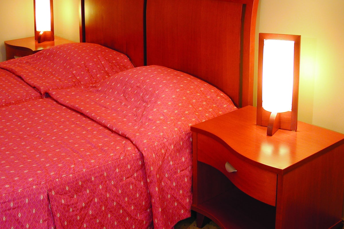 A hotel room with a wooden headboard, two bedside tables with lamps, and a red bedspread.