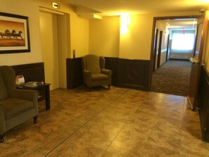 A hotel lobby with a carpeted floor, a framed picture on the wall, a chair, a small table with a red object, and a hallway leading to an exit.