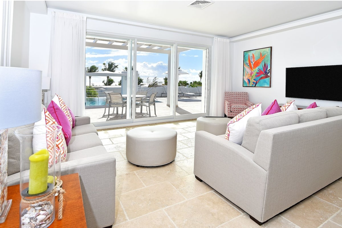 Ein modernes Wohnzimmer mit grauem Sofa, einem Couchtisch und einem Fernseher. Ein großes Fenster bietet Blick auf einen Pool und Palmen.
