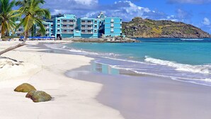 Un hotel frente a la playa con varios balcones, una playa de arena y una colina rocosa al fondo.