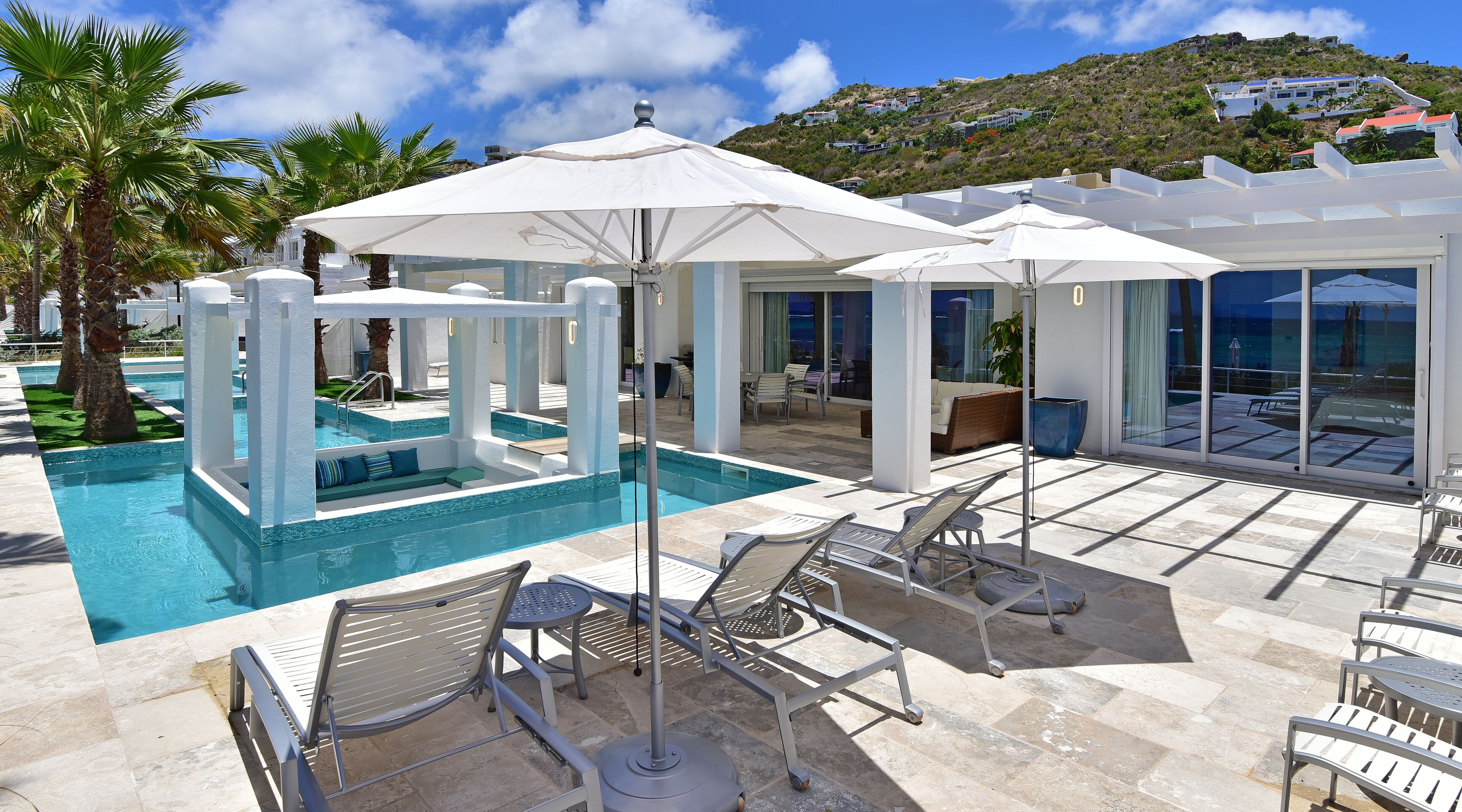 A pool area with lounge chairs and umbrellas, a glass-enclosed patio, and a view of a hillside with buildings.