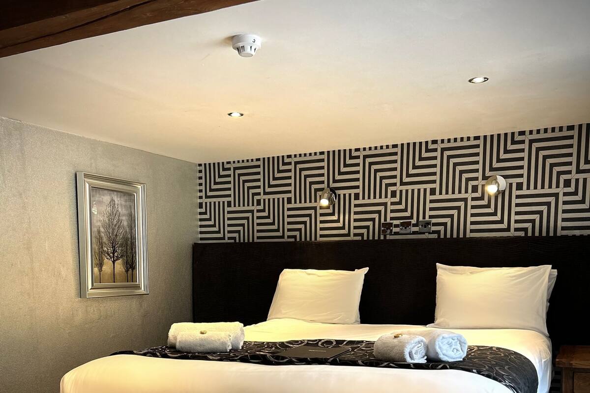 A neatly made bed with white linens and two pillows, a headboard with a geometric pattern, a framed picture on the wall, and a patterned ottoman.