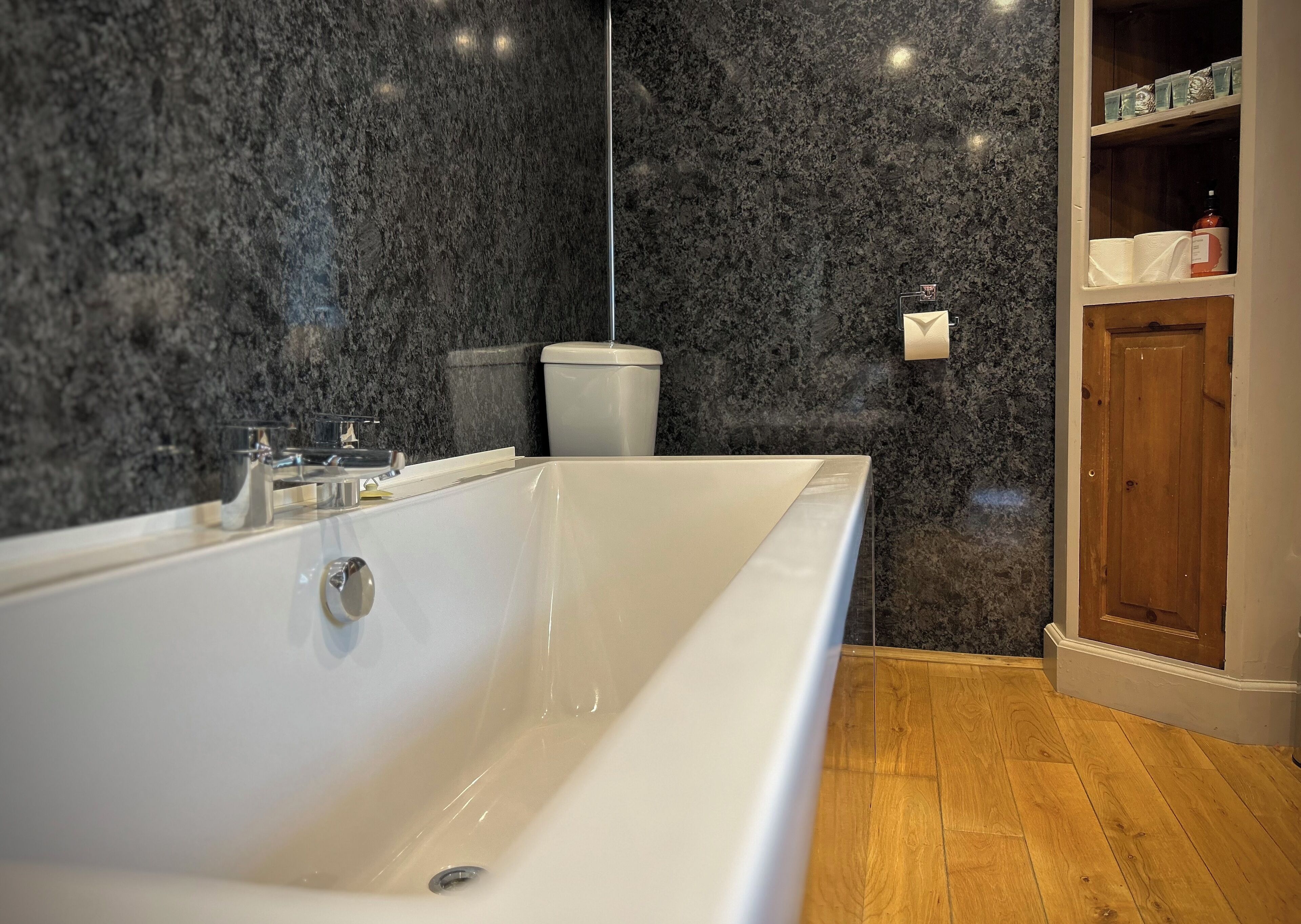 A modern bathroom with a large white bathtub, dark marble walls, and a wooden floor.