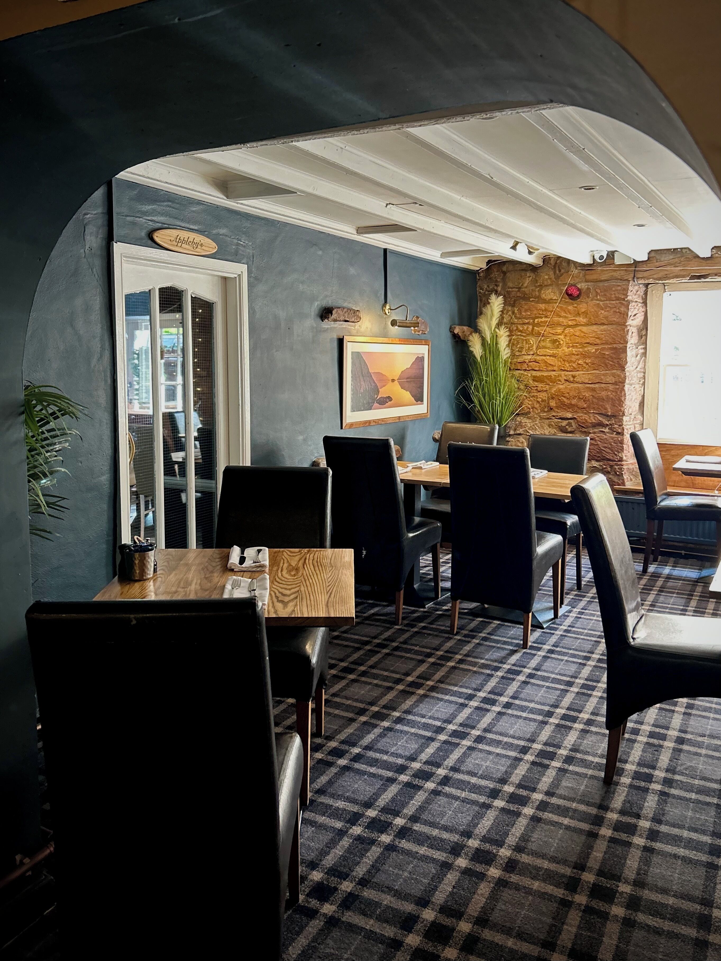 A cozy dining area with a checkered floor, wooden tables, and black chairs. A framed picture hangs on the wall, and a potted plant adds greenery.