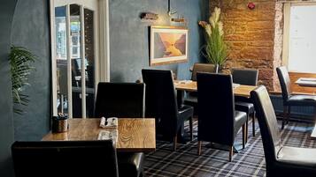 A cozy dining area with a checkered floor, wooden tables, and black chairs. A framed picture hangs on the wall, and a potted plant adds greenery.