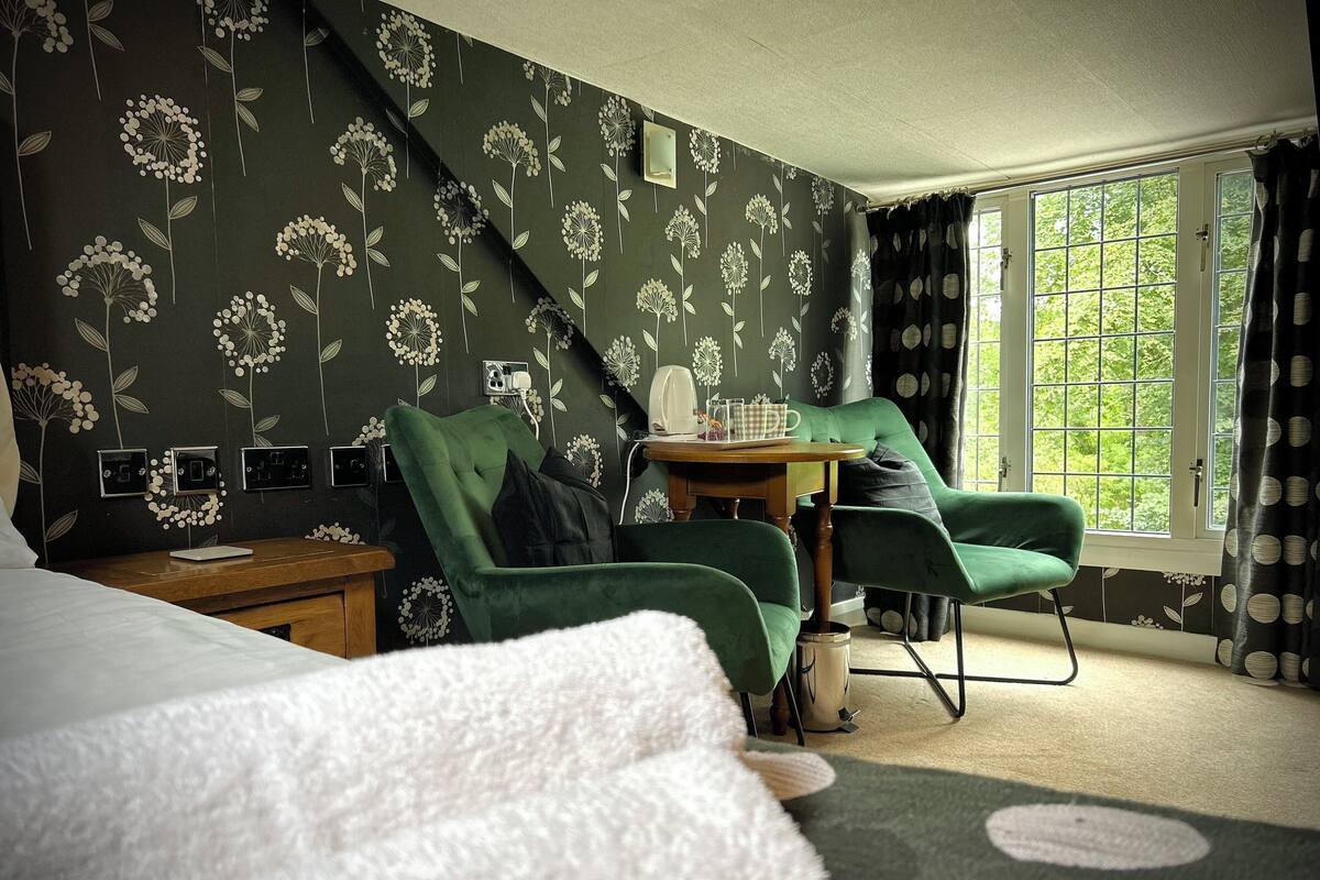 A hotel room with a bed, two green armchairs, a small table with a kettle and a cup, and a window with curtains.
