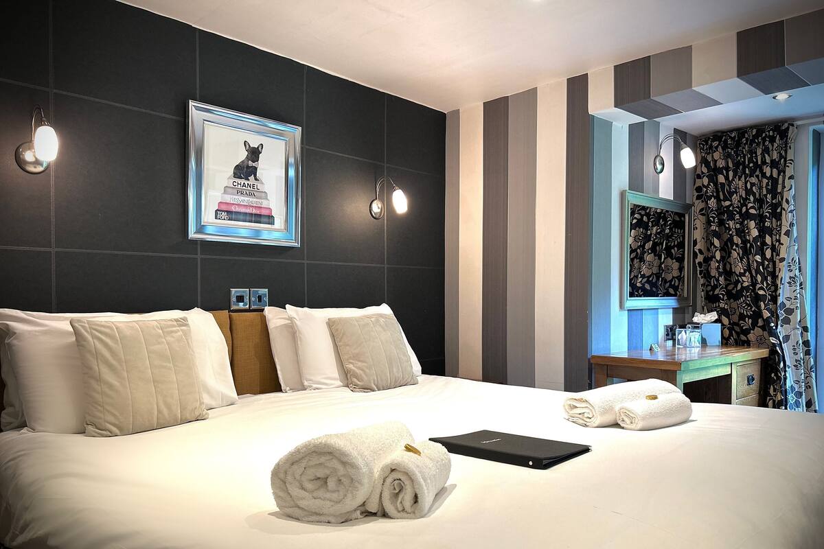 A hotel room with a large bed, a bedside table with a lamp, and a framed picture of luxury brands on the wall.