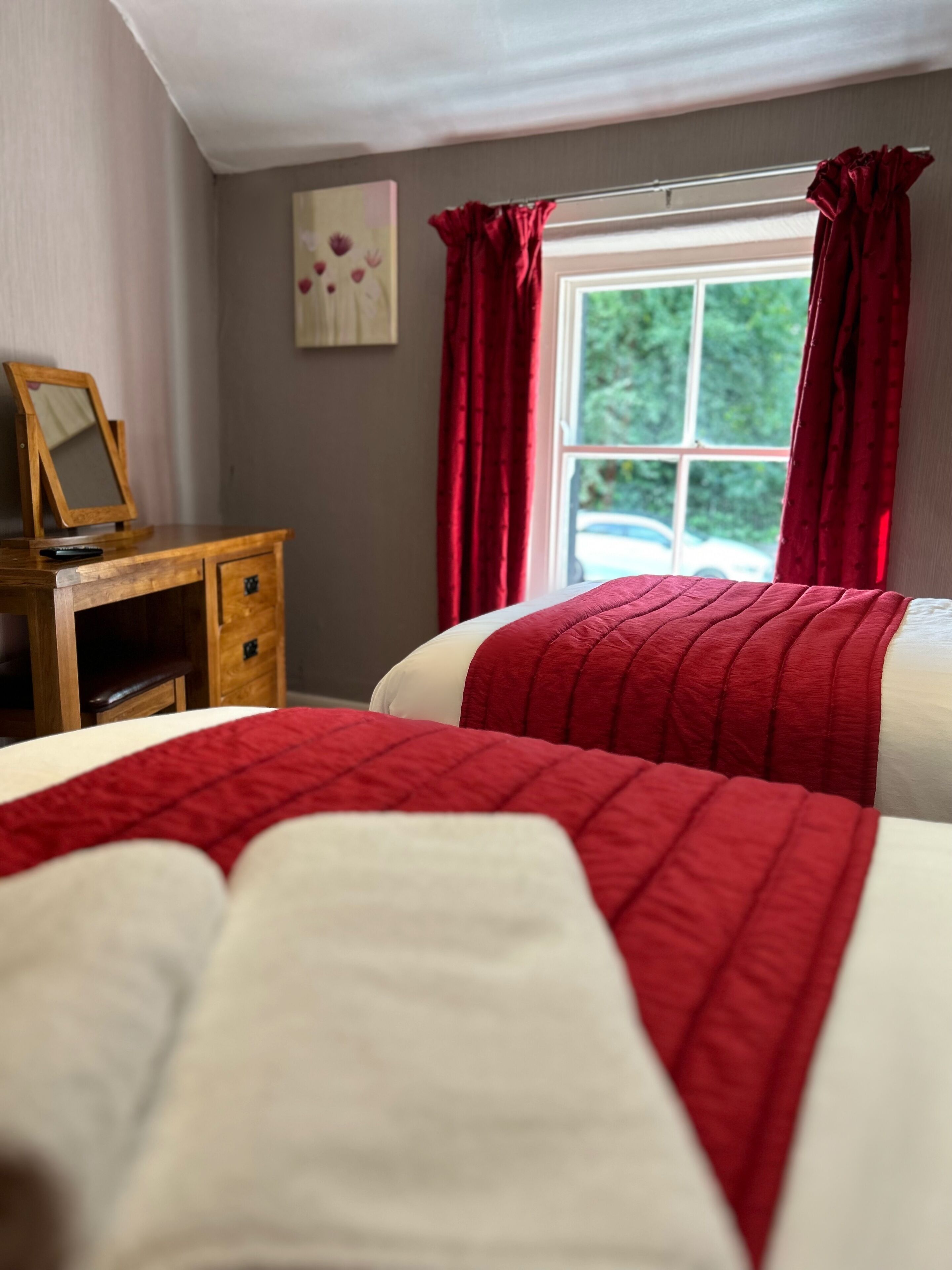 Quarto com duas camas, penteadeira de madeira, espelho e janela com cortinas vermelhas.