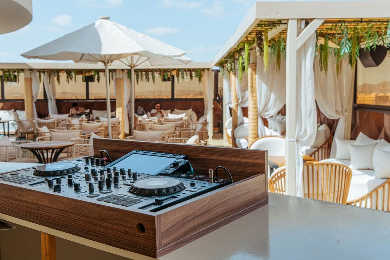 A DJ booth with a control panel, surrounded by outdoor seating with umbrellas and cushions.