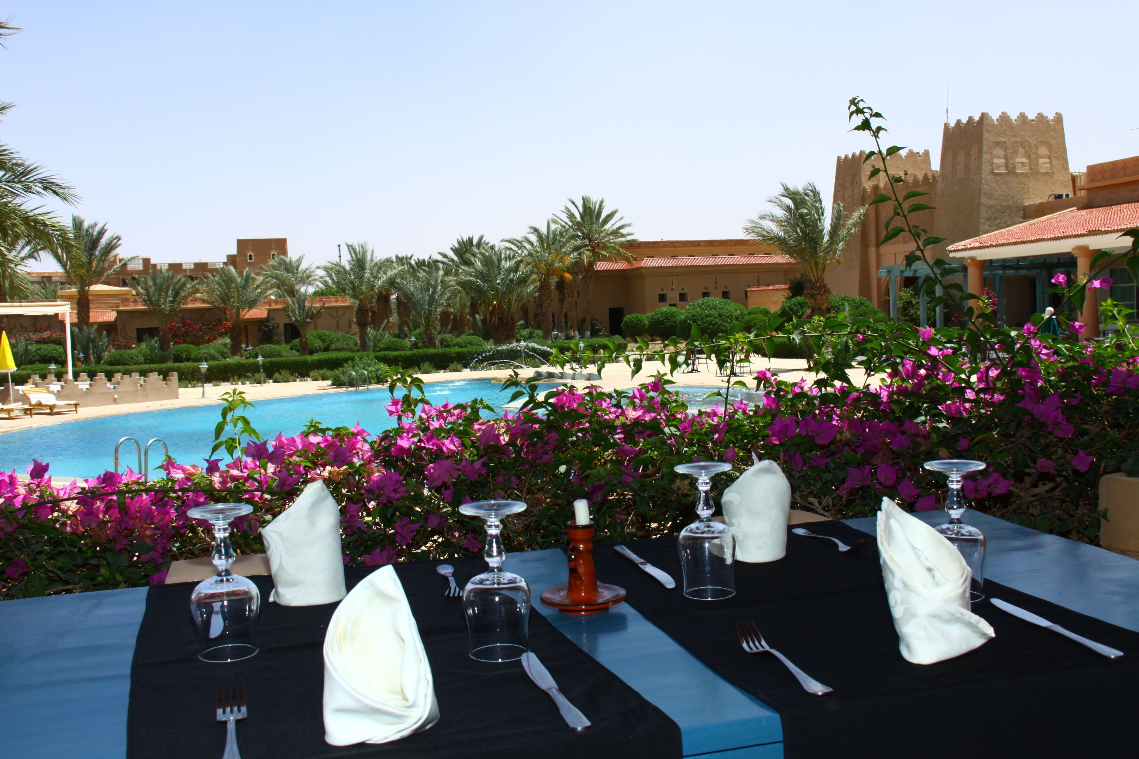 A dining table set for a meal with a view of a swimming pool and a castle-like building in the background.