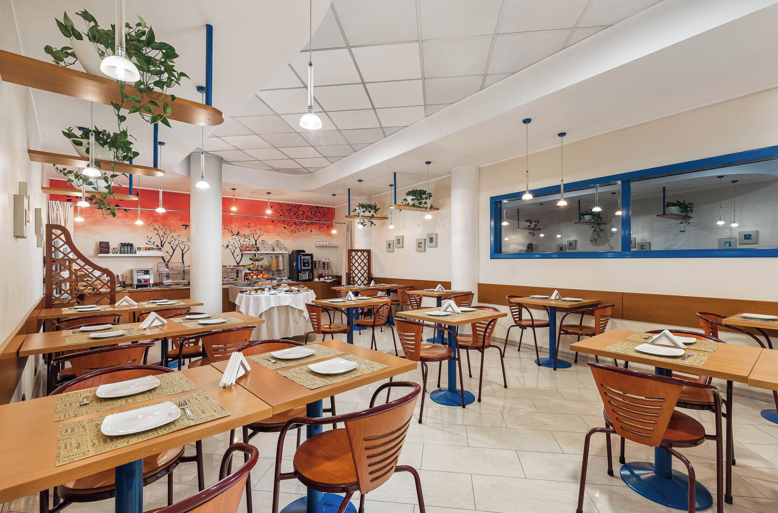 A restaurant with wooden tables and chairs, hanging plants, and a red wall with a tree mural.