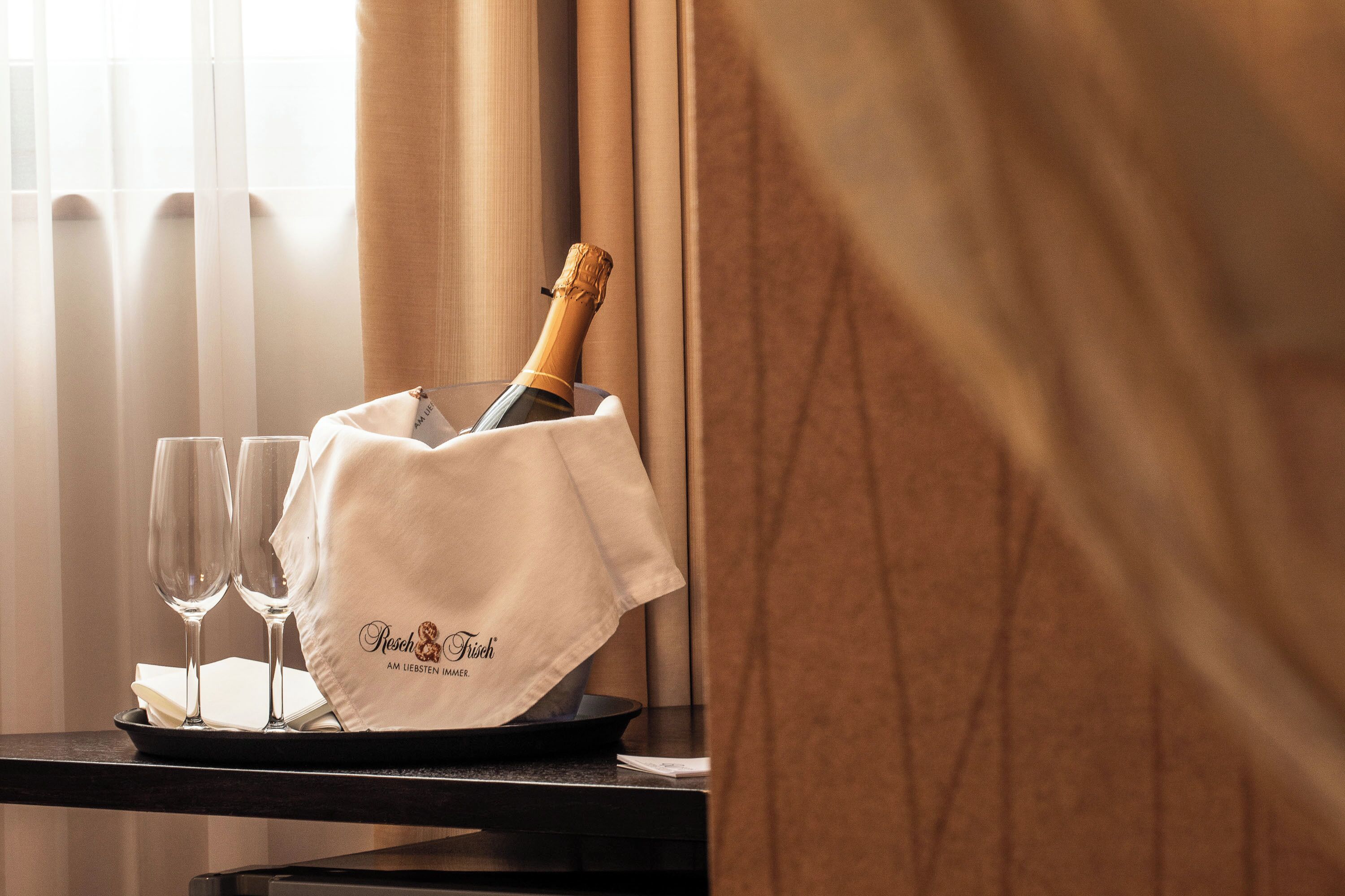 A tray with a bottle of champagne, two glasses, and a napkin on a table near a window with curtains.