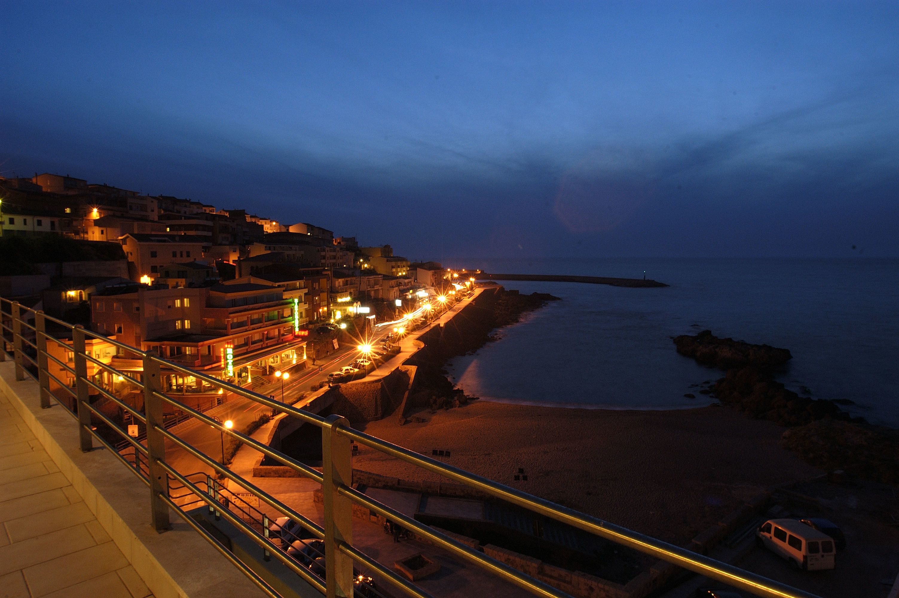 A coastal town at dusk with illuminated streets and buildings, a beach, and a pier extending into the water.