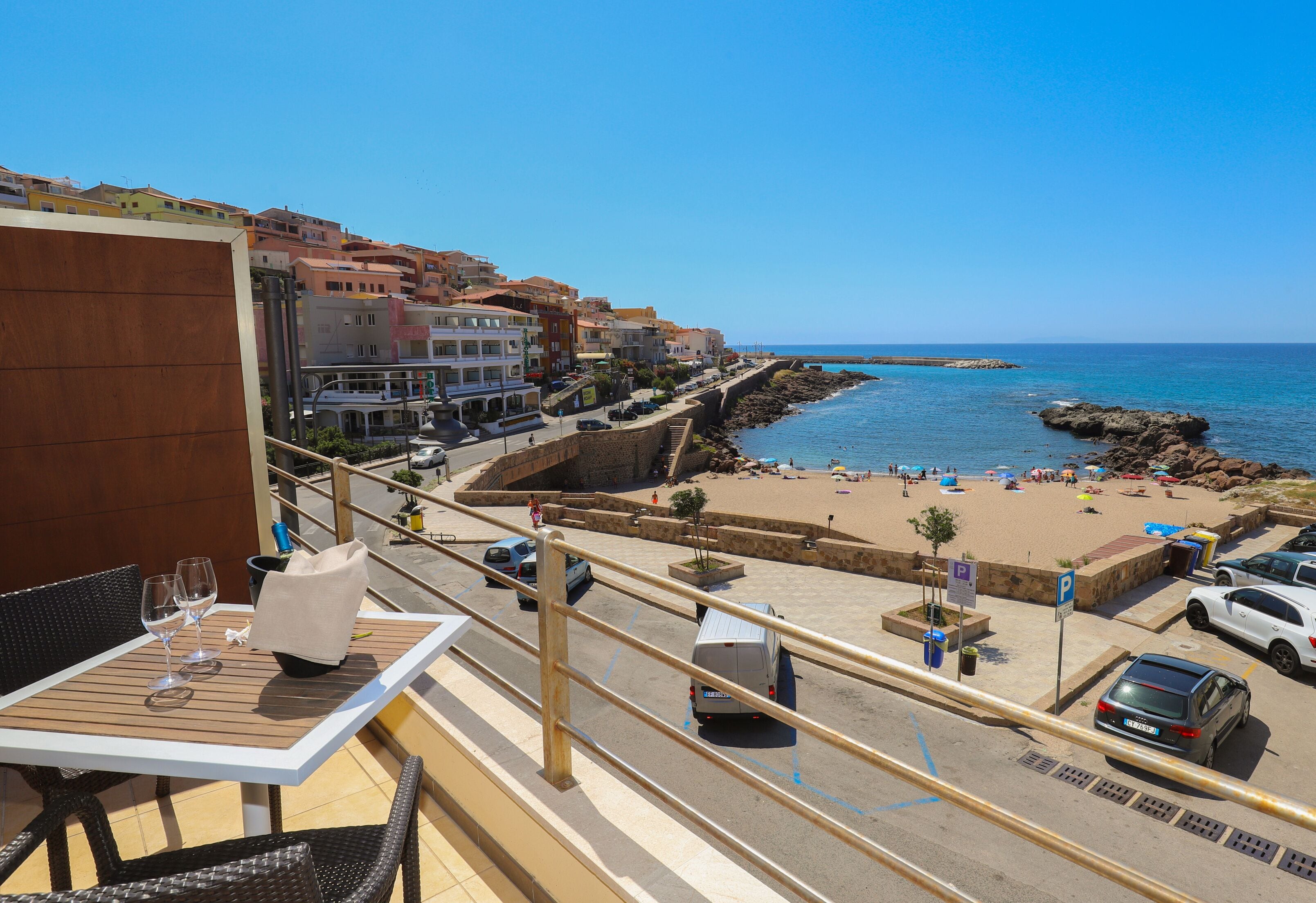 A balcony with a table and chairs, overlooking a beach with people and cars.