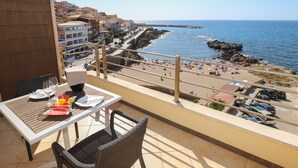 A balcony with a table set for two, overlooking a beach with people and umbrellas. - Janus Hotel (Castelsardo)
