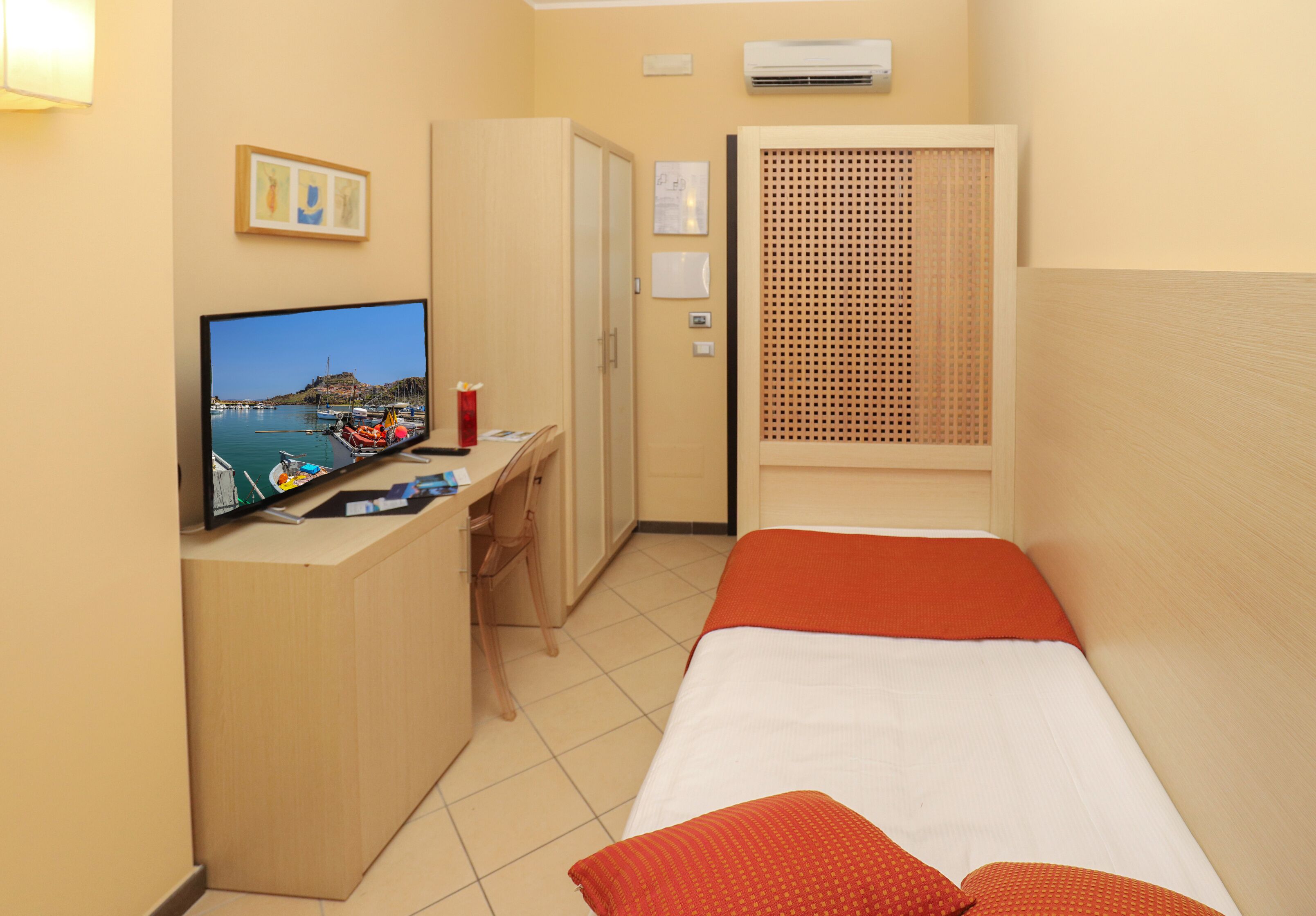 A compact hotel room with a bed, a desk with a computer, a TV displaying a scenic view, and a window with a view of boats.