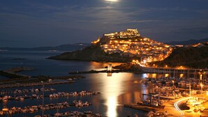 A coastal town at night with a prominent castle-like structure on a hill, a marina with boats, and a bridge connecting the town to the mainland. - Janus Hotel (Castelsardo)
