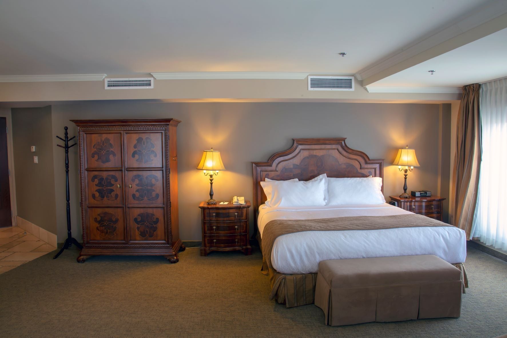 A hotel room with a large bed, two bedside tables with lamps, a wooden wardrobe, and a carpeted floor.
