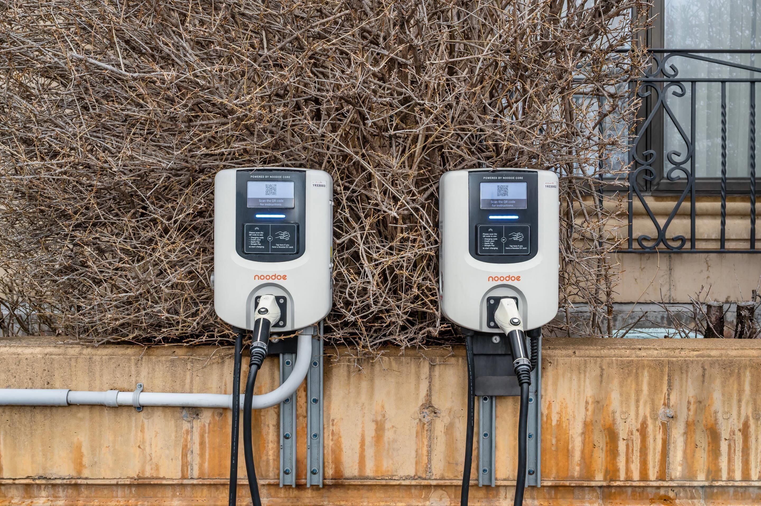 Two outdoor electrical boxes mounted on a wall with a metal railing and a bush in the background.