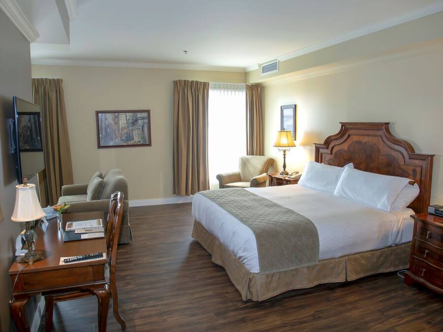 A hotel room with a large bed, a desk with a lamp, a chair, a television, and a painting on the wall.
