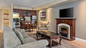 A living room with a fireplace, a television, and a kitchen in the background. - Hotel Brossard, an Ascend Collection Hotel (Brossard)