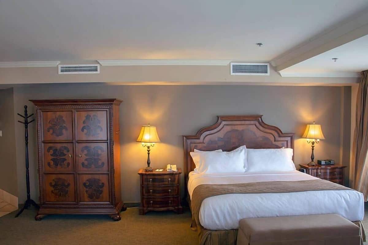 A hotel room with a large bed, two bedside tables with lamps, a wooden cabinet, and a ceiling vent.