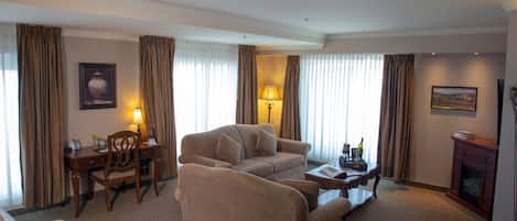 Une chambre d’hôtel avec un canapé beige, un bureau en bois, une lampe, une cheminée et un tableau au mur.