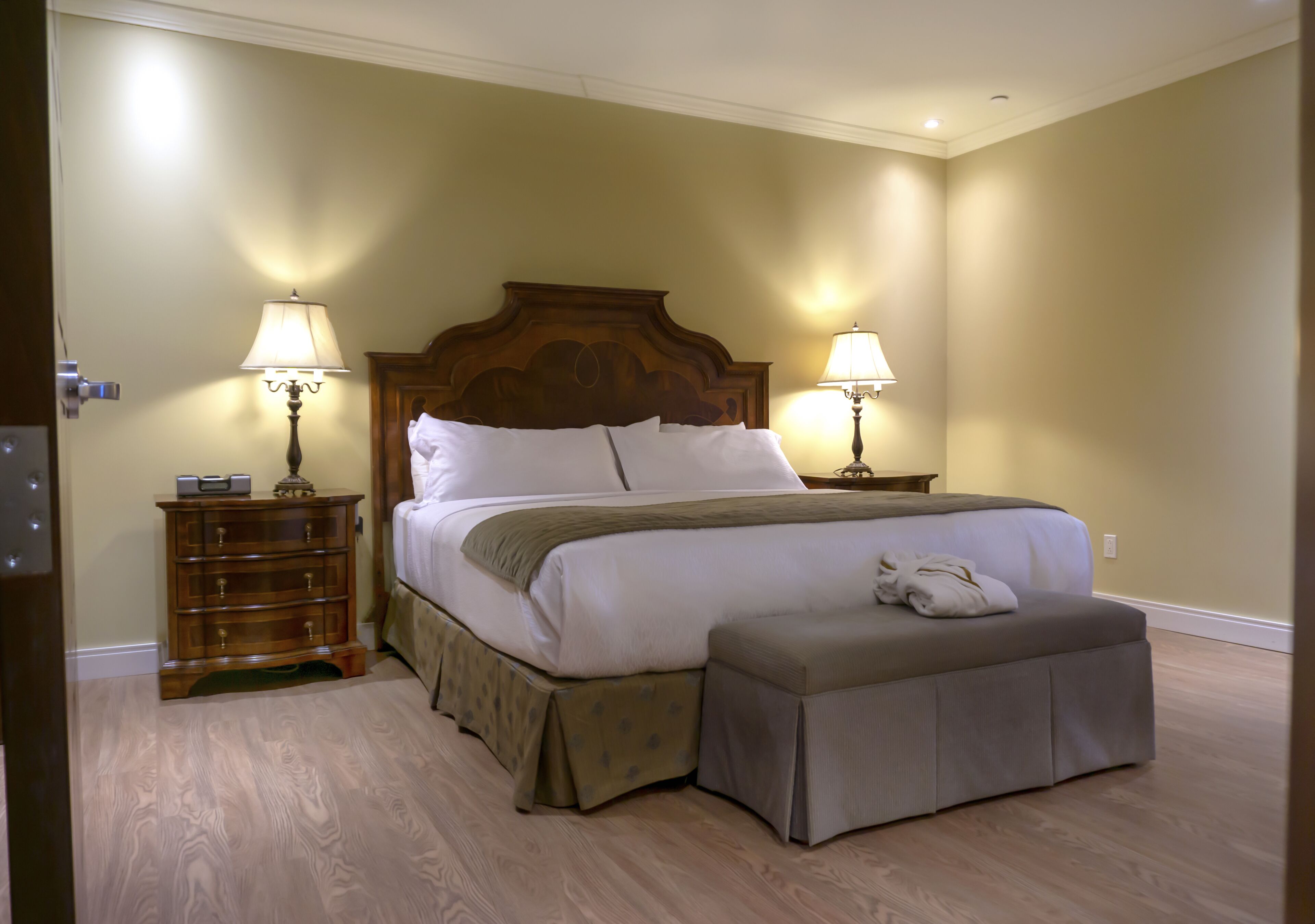 Una cama bien hecha con cabecera de madera, dos mesitas de noche con lámparas, un banco al pie de la cama y un aparador de madera.