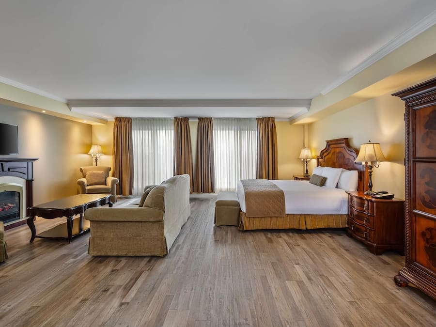 A hotel room with a bed, sofa, armchair, wooden floor, fireplace, and a TV.