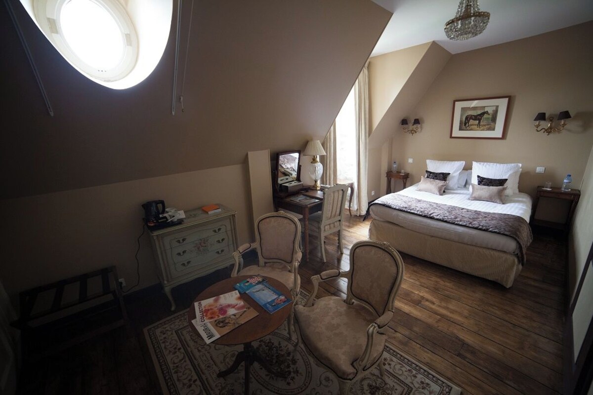 A cozy bedroom with a bed, bedside tables, a desk, and a chair. There is a window with curtains, a chandelier, and a painting on the wall.