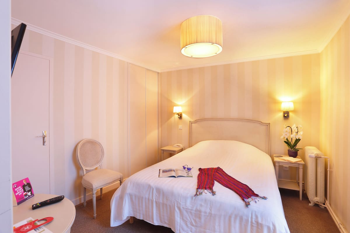 Une chambre d’hôtel avec un grand lit, une chaise, une table de chevet avec une lampe, une petite table avec une plante et un radiateur.