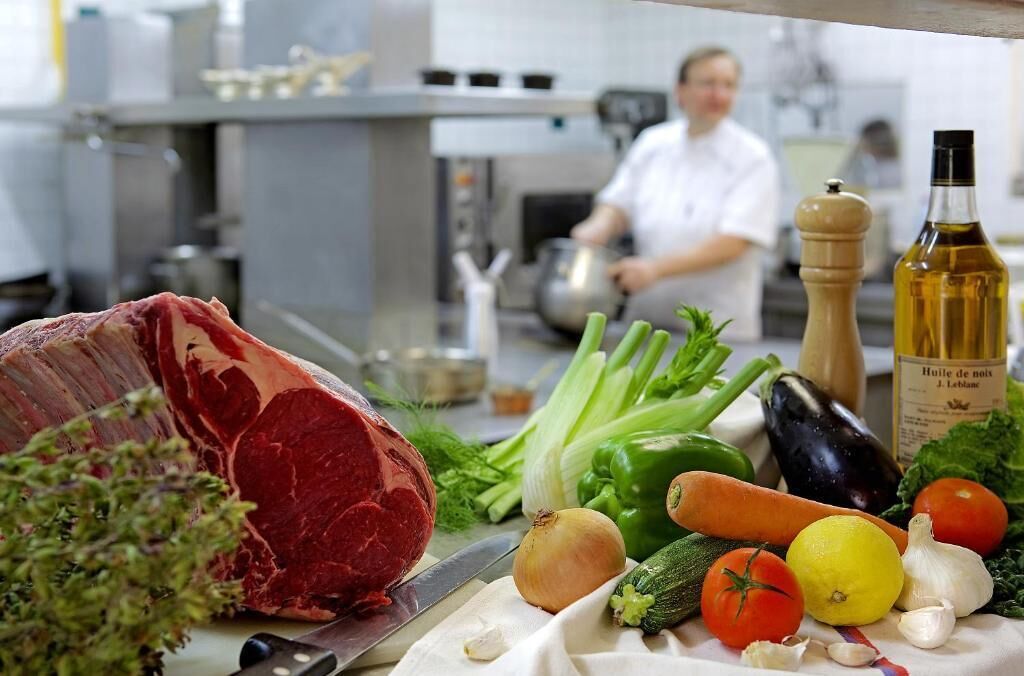 A kitchen counter with a large piece of meat, vegetables, and a bottle of oil.