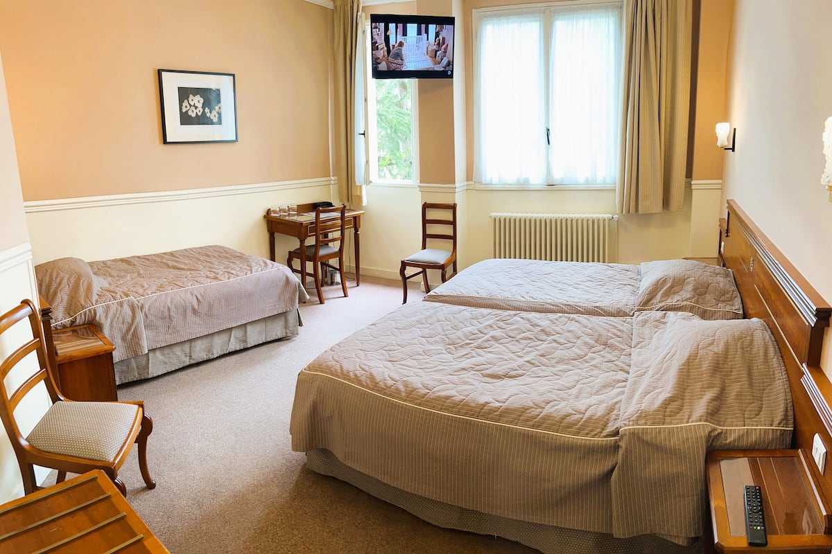 Une chambre d’hôtel avec deux lits simples, une chaise en bois, une petite table, un téléviseur et une fenêtre avec des rideaux.