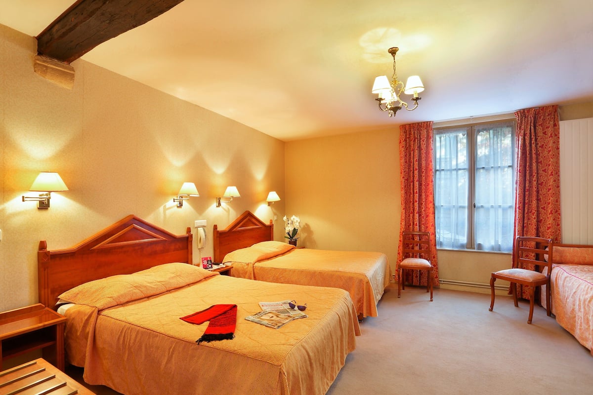 Une chambre d’hôtel avec deux lits, une tête de lit en bois, un lustre, une fenêtre avec des rideaux et une chaise en bois.