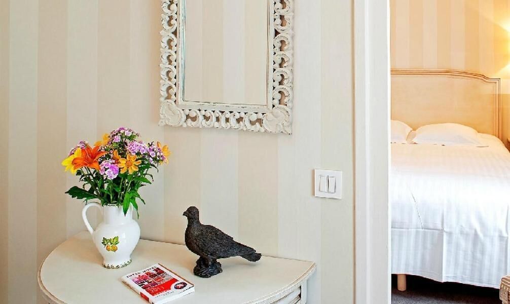 A room with a bed, a white wall with a decorative mirror, a small table with a vase of flowers, and a pigeon statue.