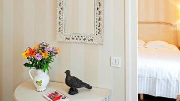 A room with a bed, a white wall with a decorative mirror, a small table with a vase of flowers, and a pigeon statue.