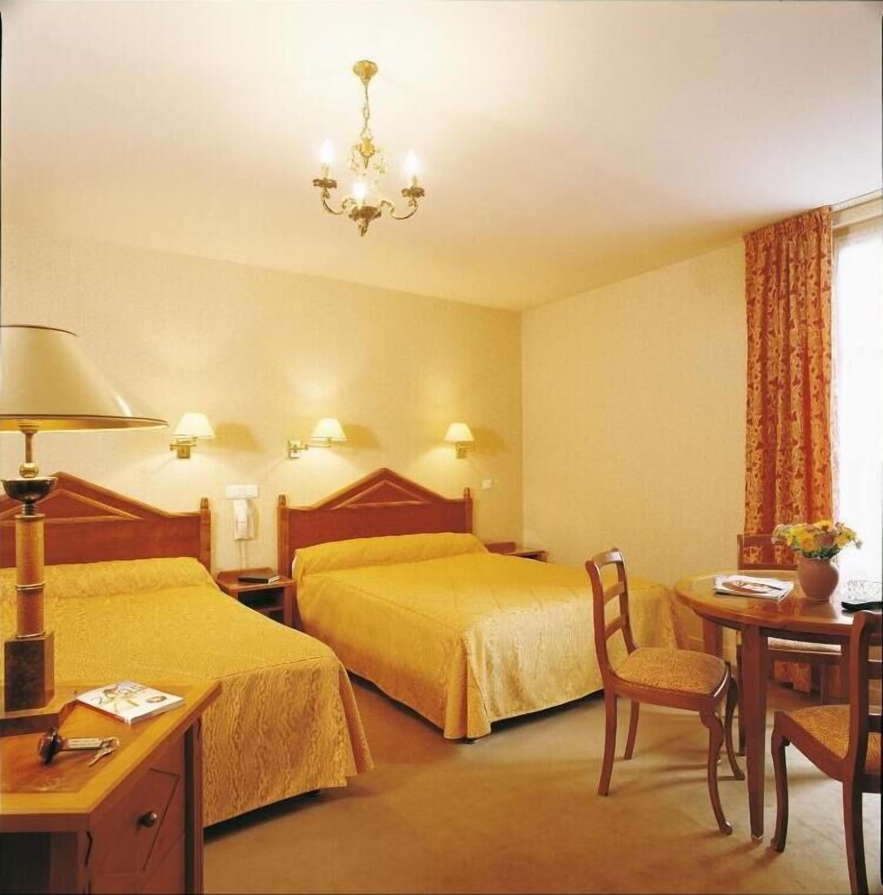 A hotel room with two beds, a desk, a chair, a table, and a chandelier.
