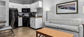 A compact kitchen with white cabinets, a black refrigerator, and a microwave. There is a grey sofa and a wooden table in the living area. - Club Wyndham at Majestic Sun (Miramar Beach)