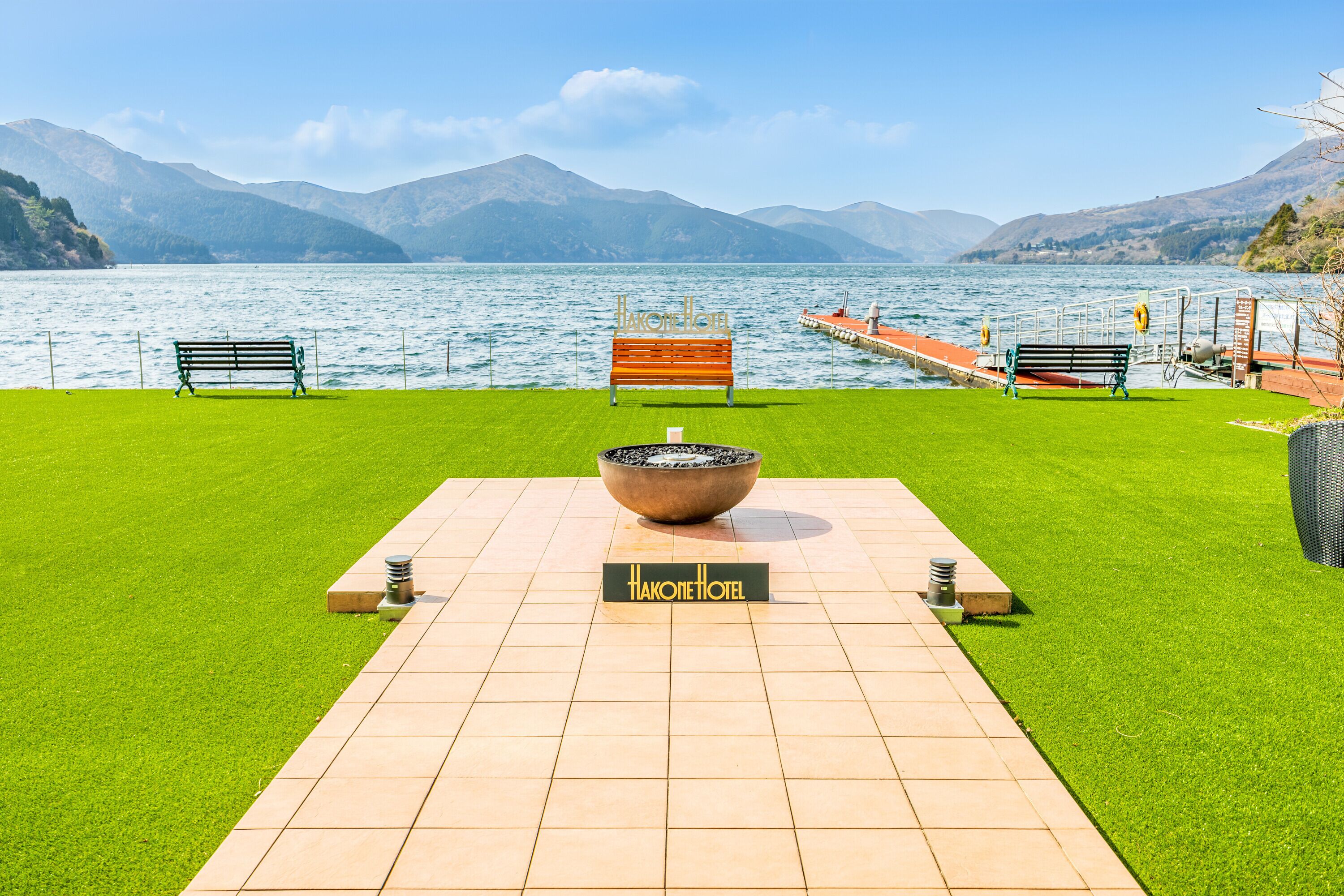 Blick auf einen See mit einem Schild für das Hakone Hotel, einer hölzernen Bank und einem steinernen Becken.