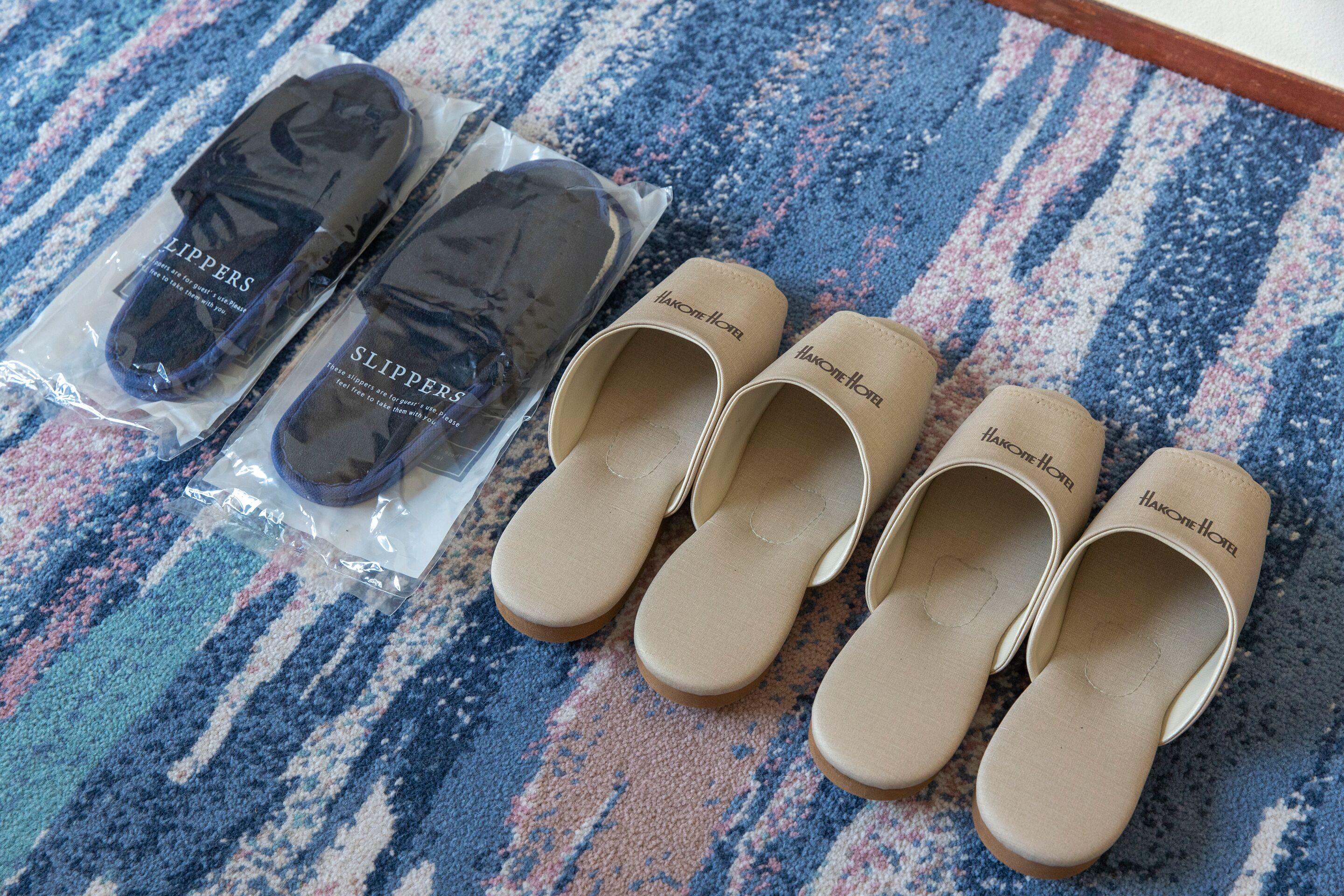 Un par de pantuflas negras y beige con el logotipo de "Hakon Hotel" sobre una alfombra a rayas.