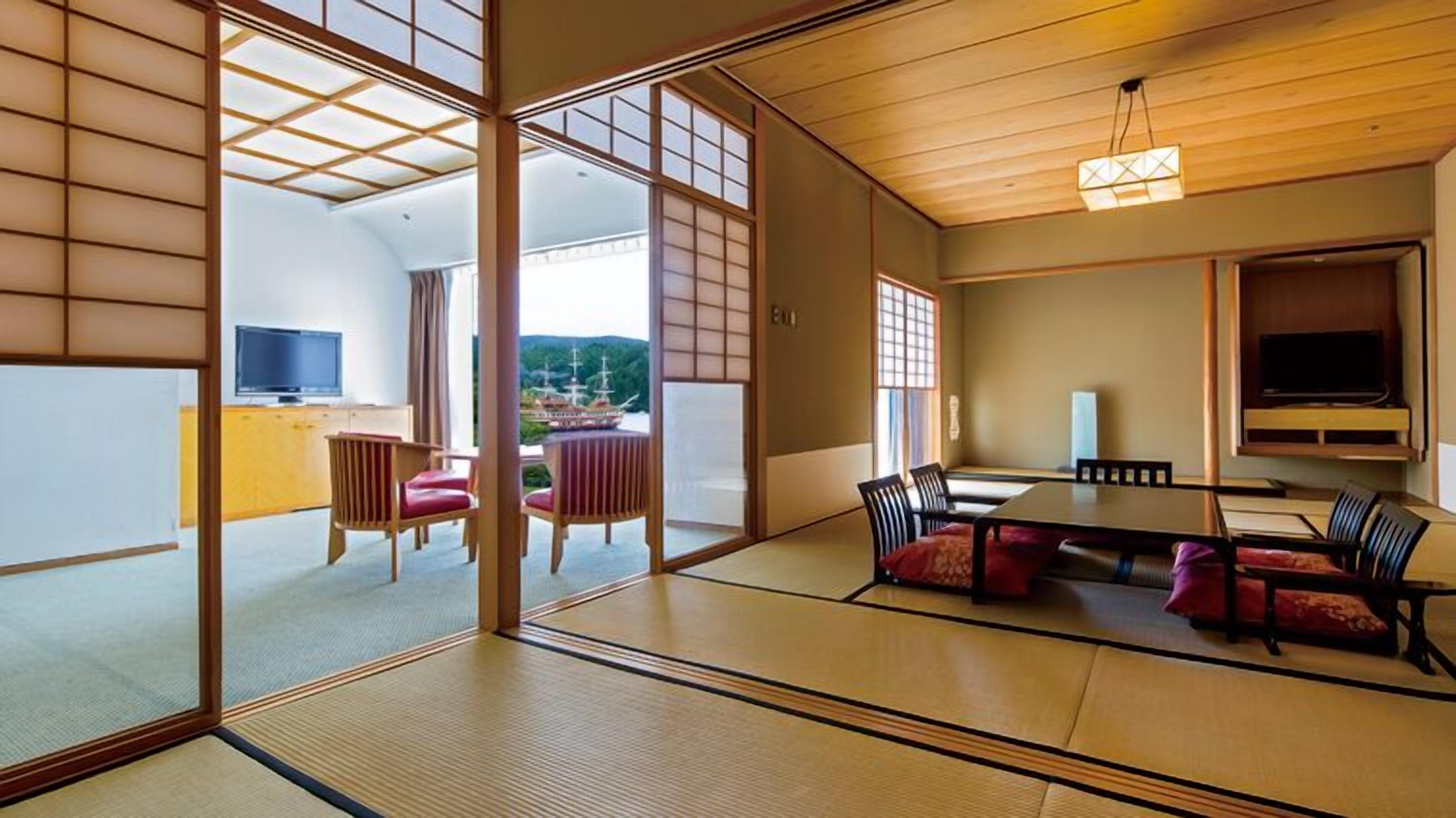 A traditional Japanese room with tatami flooring, a low table with cushions, a television, and a view of a traditional building.