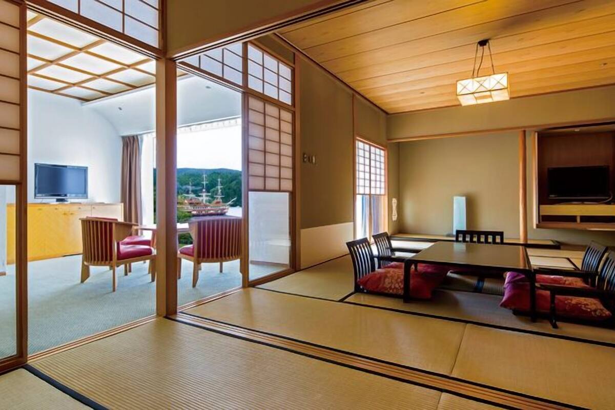 Et traditionelt japansk rum med tatamimåtter, et lavt bord med puder, et fjernsyn og udsigt til en traditionel bygning.