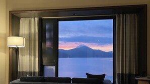 Habitación de hotel con una cama grande, un sofá, un escritorio y vistas a una montaña y un lago al atardecer.
