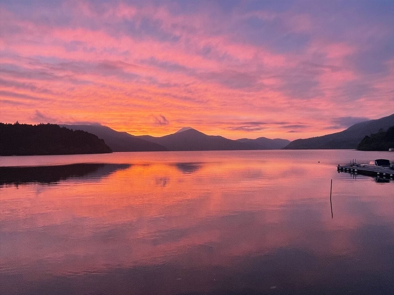 A calm lake at sunset with mountains in the background and a dock with boats.