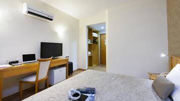 A hotel room with a bed, a desk with a laptop, a television, a mini-fridge, and an air conditioner.