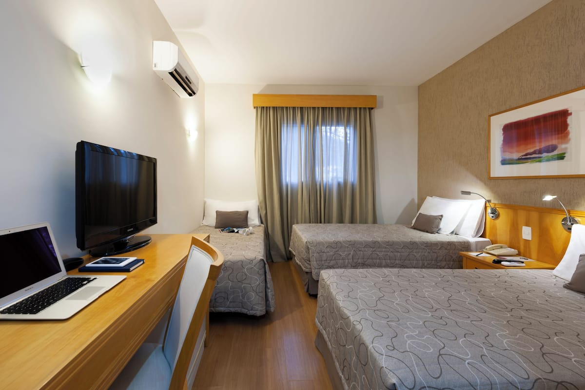 A hotel room with a desk, a flat-screen TV, two beds, a window with curtains, a painting on the wall, and a bedside lamp.