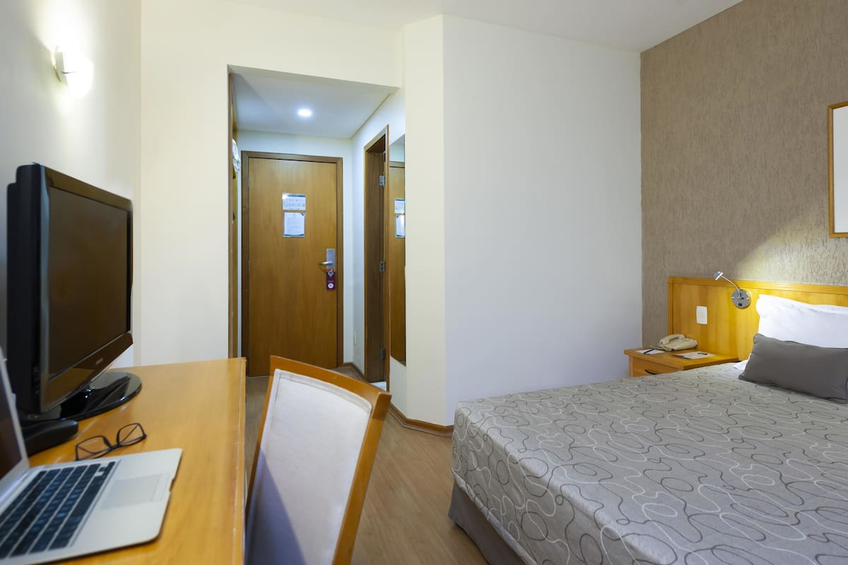 A hotel room with a bed, a desk with a laptop, a chair, a bedside table with a phone, and a door with a sign.